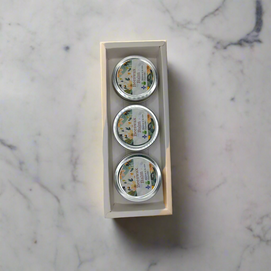 Three small round tins from Mielbrut.com, featuring colorful floral labels, are neatly arranged in a cream box on a marble surface. This 3 pack beeswax balms set promises natural benefits and skin hydration. The background is light with grey veins.