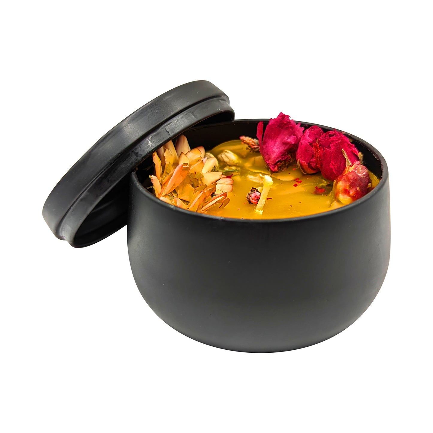 A small, black, round container with its lid partially open is filled with a yellow substance, reminiscent of a Handcrafted Beeswax Candle with Wildflowers in Sleek Black Tin by Mielbrut.com, topped with wildflowers and herbs. The container has a matte finish, and the dried flowers include red rose petals and other natural elements.