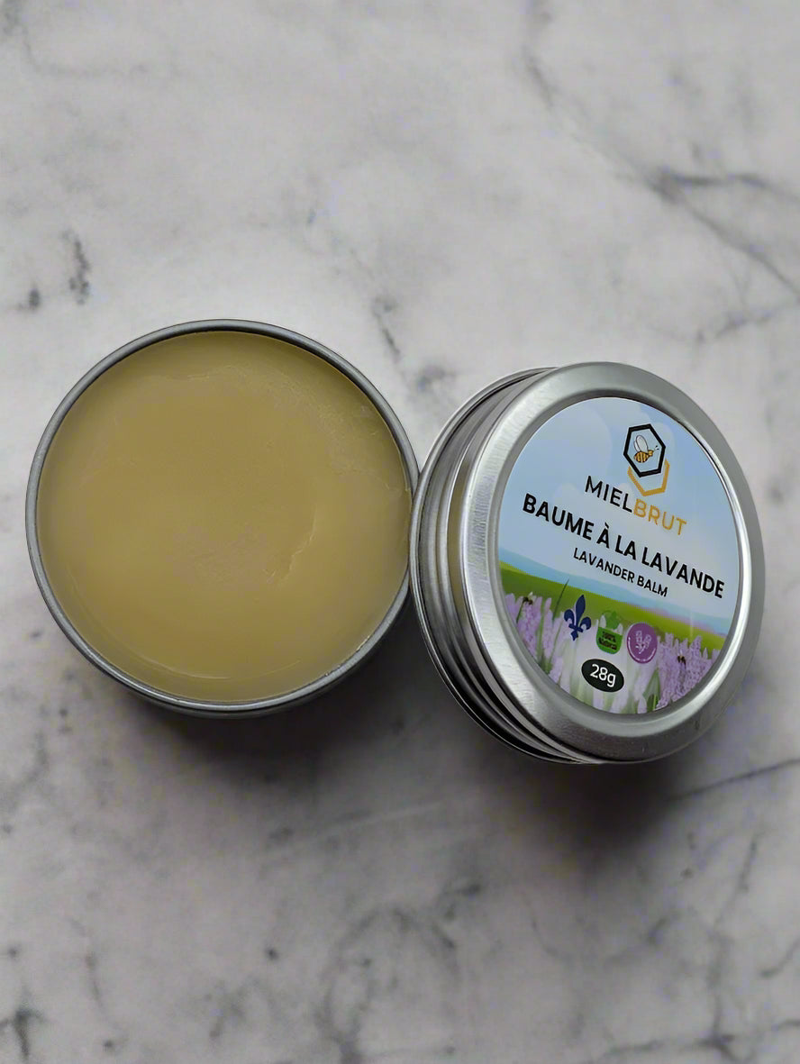 A 1oz tin of Mielbrut.com's Lavender Beeswax Balm sits open, revealing its creamy, solid balm made from natural ingredients. The nearby lid displays lavender illustrations on a light label. Ideal for soothing dry skin, this balm rests elegantly on a marble surface.