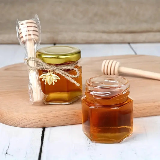 Two small hexagonal glass jars filled with premium quality honey from Mielbrut.com are placed on a wooden cutting board. One jar is sealed with a golden lid and decorated with a honey dipper tied with a string, while the other jar is open with a honey dipper lying nearby, showcasing the customizable packaging offered by our Honey Jar Filling Service: Perfect for Weddings and Corporate Events.