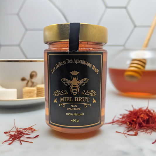 A jar of "Mielbrut.com" raw honey labeled "Saffron Elixir: Pure Quebec Honey Infused with the Golden Essence of Iranian Saffron," non-pasteurized and 100% natural, rests on a countertop. In the background are Iranian saffron threads, a honey dipper in a bowl of saffron honey, and a plate with sugar cubes. The jar is 450 grams.