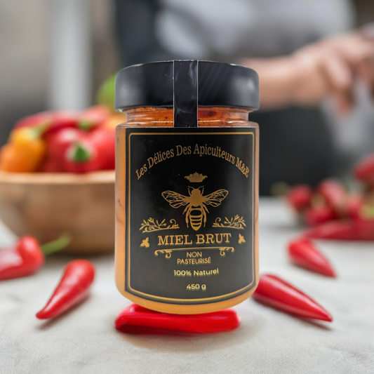 A jar of "Zesty Honey Infused with Heat: Spice up Your Palate!" with a black label featuring a bee and the French text "Mielbrut.com" is placed on a table. Surrounding the jar are red chili peppers, hinting at sweet and spicy culinary adventures. There's a blurred bowl of strawberries in the background.
