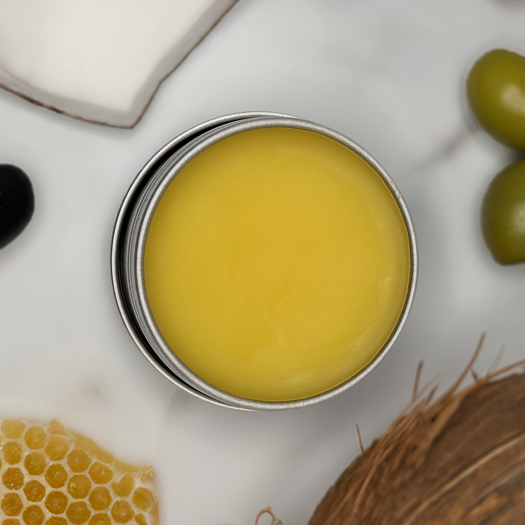 A round tin container filled with solid yellow balm is placed on a white surface. Surrounding it are various natural ingredients: a piece of coconut, honeycomb, olives, and what appears to be a slice of black olive. The image suggests the Mielbrut.com's Beeswax Balm: Moisturizer for lips, hands, feet and skin is honey-infused for natural skincare excellence.