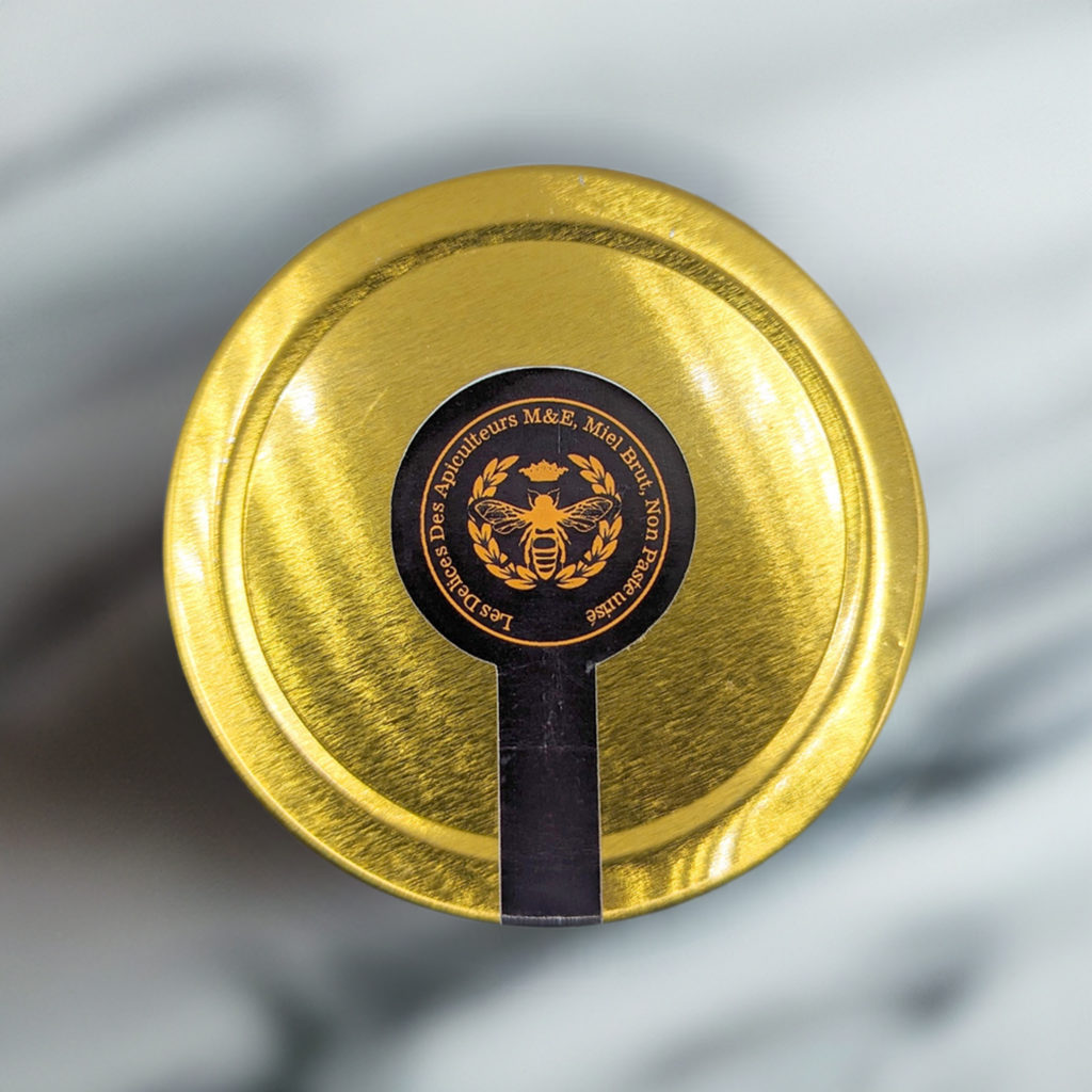 A top-down view of a golden-colored jar lid with a round, black label in the center. The label features an illustration of a bee, surrounded by the text "Mielbrut.com Miel Pur," reflecting their beekeeping passion. The background appears to be a blurry grey marble surface, hinting at this Beekeeper's Special Reserve: Exceptional Catalpa Honey's limited edition allure.