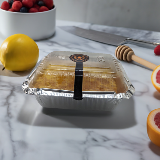 A foil container with a transparent plastic cover and a black seal is placed on a marble surface. Surrounding it are various fruits: a yellow lemon, half of a grapefruit, and a white bowl with mixed berries. A large knife, Quebec Gold: Pure Honey Comb Straight from Nature's Hive by Mielbrut.com, and a honey dipper loaded with raw honey are also nearby.