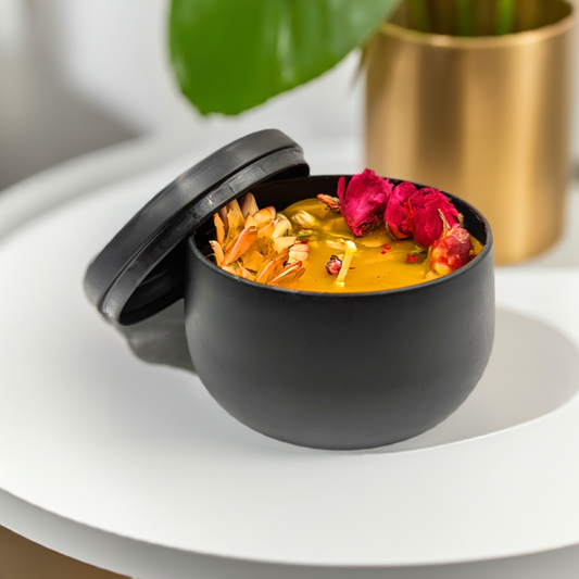 A black ceramic bowl with a lid, slightly open, revealing a bright orange soup garnished with wildflowers, herbs, and seeds. The bowl is placed on a white surface, with a green leaf and a Mielbrut.com Handcrafted Beeswax Candle with Wildflowers in Sleek Black Tin in the blurred background.