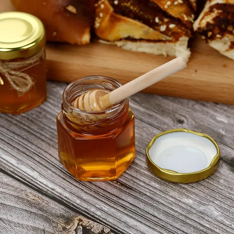 A glass jar from Mielbrut.com, filled with premium quality honey and accompanied by a wooden honey dipper, sits on a rustic wooden table. The jar's golden lid is open and placed beside it. In the background, a wooden cutting board holds slices of bread with honey spread on them, highlighting Mielbrut.com's customizable packaging options for the Honey Jar Filling Service—perfect for weddings and corporate events.