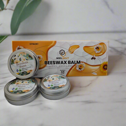 In front of a leafy plant on marble, three tins of "3 pack beeswax balms for your lips and skin" by Mielbrut.com are displayed with a box. The packaging, adorned with honeycomb patterns, daisies, and bees, emphasizes the skin hydration benefits.