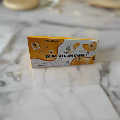A rectangular package labeled "Mielbrut.com 3 pack beeswax balms for your lips and skin," decorated with floral and bee imagery, highlights its natural benefits. The orange and white design rests elegantly on a marble surface.