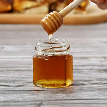 A small glass jar filled with premium quality honey from Mielbrut.com sits on a wooden surface. Above the jar, a honey dipper drips honey into the Honey Jar Filling Service—perfect for weddings and corporate events. The background is blurred, drawing attention to the custom honey jar filling and dipper.