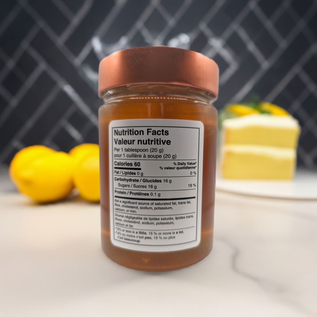 A glass jar of Mielbrut.com Beekeeper's Amber Wildflower Honey with a copper lid features a bilingual Nutrition Facts label. Blurred lemons and a dessert rest on a kitchen counter with a black tiled backsplash in the background.