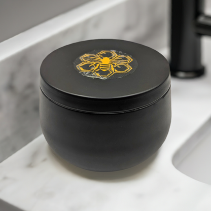 A matte black container with a lid, decorated with a yellow bee emblem, houses the Handcrafted Beeswax Candle with Wildflowers in Sleek Black Tin by Mielbrut.com. It is placed on a white marble countertop next to a black faucet.