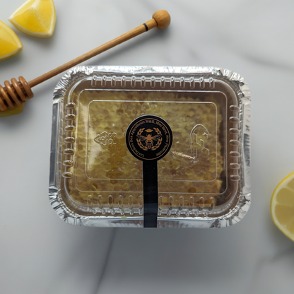 A sealed rectangular container of Quebec Gold: Pure Honey Comb Straight from Nature's Hive by Mielbrut.com, with a black and gold label, rests on a marble surface. Surrounding the container are lemon wedges and a wooden honey dipper. The packaging is clear on top, displaying the exquisite honeycomb inside.