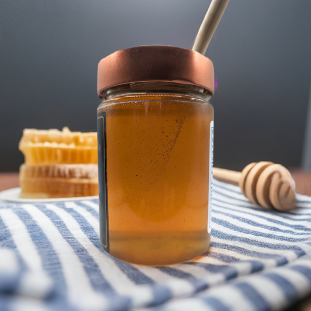 A jar of Mielbrut.com Beekeeper's Amber Wildflower Honey with a copper lid and honey dipper sits on a striped cloth, while pieces of honeycomb appear on a white plate in the background.