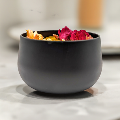 A sleek black ceramic bowl holds a small assortment of wildflowers, including yellow, orange, and red petals, sitting on a smooth, light-colored surface. The background is softly blurred, highlighting the bowl and its contents alongside a Mielbrut.com Handcrafted Beeswax Candle with Wildflowers in Sleek Black Tin.