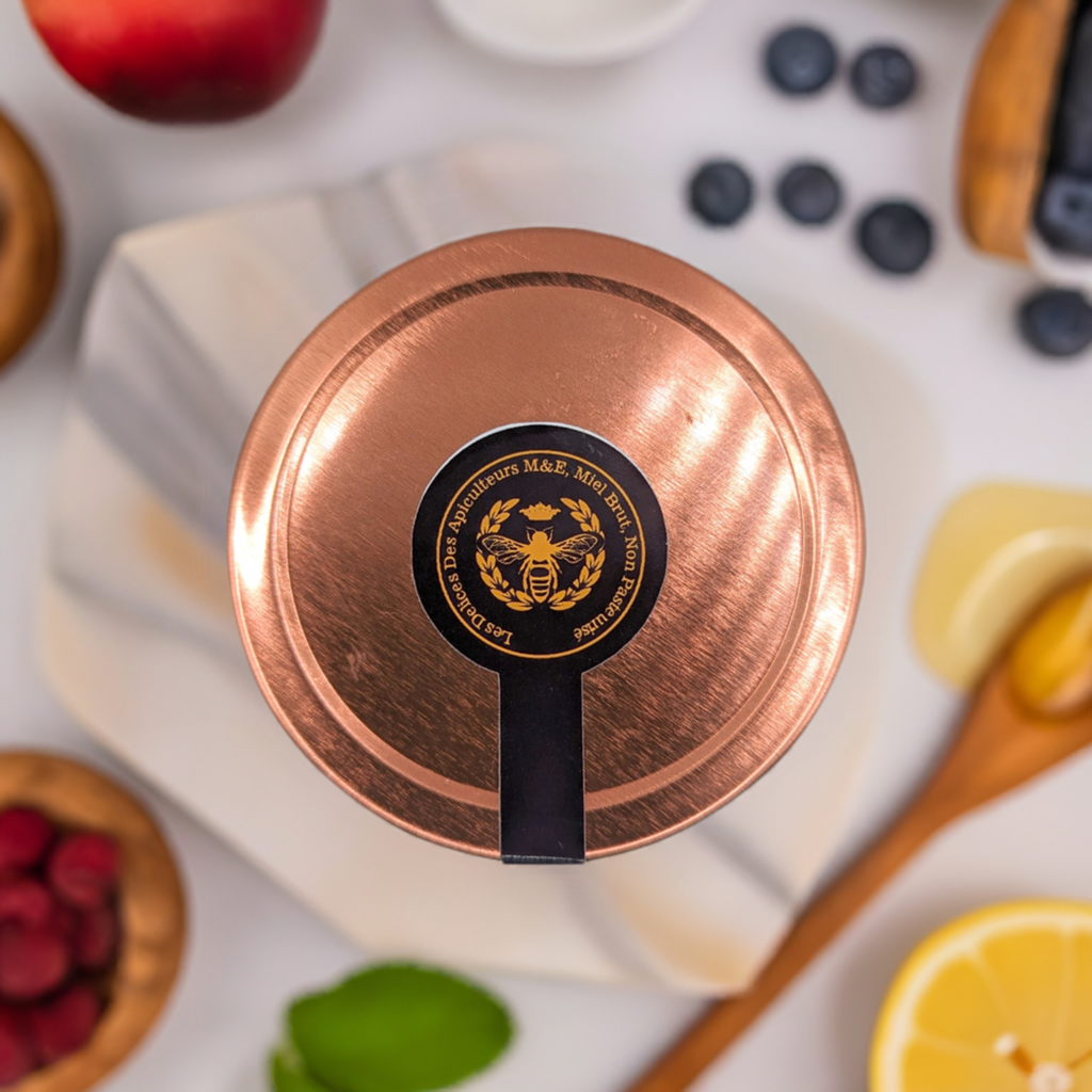A copper tin of Beekeeper's Amber Wildflower Honey by Mielbrut.com sits on marble, surrounded by blueberries, raspberries, lemon slices, an apple, a honey dipper, and green leaves—highlighting its pure Quebec honey and vibrant ingredients.