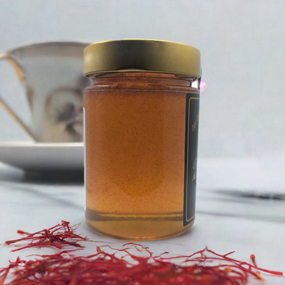 A glass jar of Saffron Elixir by Mielbrut.com—pure Quebec honey infused with the golden essence of Iranian saffron—sealed with a gold lid. Iranian saffron threads and a blurred teacup and saucer appear in the background.