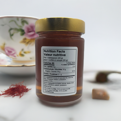 Saffron Elixir: Pure Quebec Honey Infused with the Golden Essence of Iranian Saffron by Mielbrut.com sits in a glass jar with a gold lid, nutrition label visible. A floral teacup and saucer and Iranian saffron threads are nearby on a white surface.