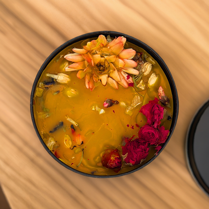A Handcrafted Beeswax Candle with Wildflowers in Sleek Black Tin from Mielbrut.com contains a vibrant yellow-orange liquid, topped with various colorful wildflowers and petals, including a large orange flower and several red petals. The handcrafted bowl is resting on a wooden surface with a blurred pattern in the background.