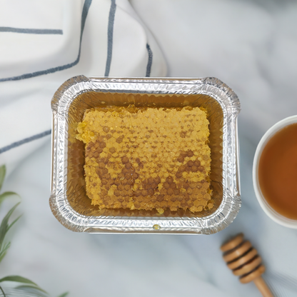 A close-up image of a Quebec Gold: Pure Honey Comb Straight from Nature's Hive by Mielbrut.com placed in a rectangular, silver-colored foil container. The container is on a marble surface with a white napkin and a cup of tea nearby. A honey dipper and a green plant are partially visible in the background, showcasing the natural beauty of raw honey.