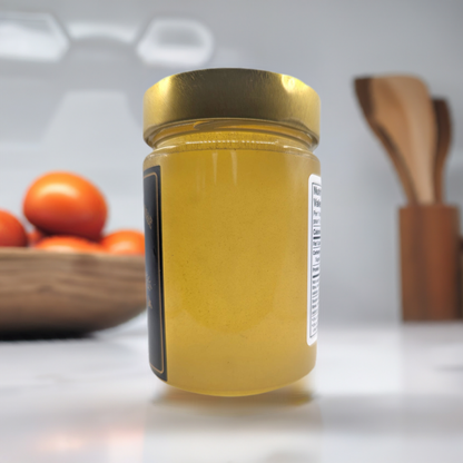 A glass jar filled with Beekeeper's Special Reserve: Exceptional Catalpa Honey from Mielbrut.com, topped with a gold lid, sits on a white kitchen counter. In the background, there is a wooden bowl containing tomatoes and wooden utensils, creating a warm and inviting kitchen atmosphere.