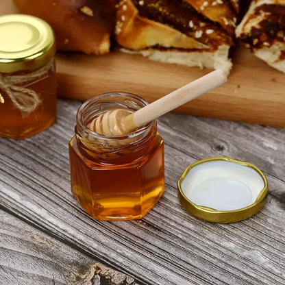 A glass jar from Mielbrut.com, filled with premium quality honey and accompanied by a wooden honey dipper, sits on a rustic wooden table. The jar's golden lid is open and placed beside it. In the background, a wooden cutting board holds slices of bread with honey spread on them, highlighting Mielbrut.com's customizable packaging options for the Honey Jar Filling Service—perfect for weddings and corporate events.