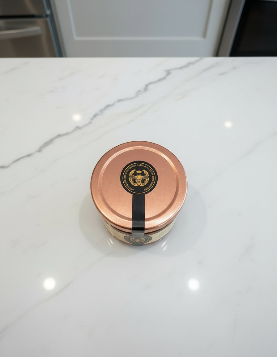 A small jar of Mielbrut.com Whipped Honey with a copper lid and black-and-gold seal sits on a white marble countertop with grey veining, against a backdrop of kitchen cabinets and appliances.